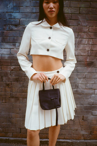 A model wears a bone colored skort and a Cropped tailored jacket in Bone mid weight suiting with long bows at back, chest pockets, and button closure against a brick wall