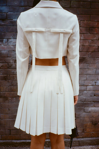 A model wears a bone colored skort and a Cropped tailored jacket in Bone mid weight suiting with long bows at back, chest pockets, and button closure against a brick wall