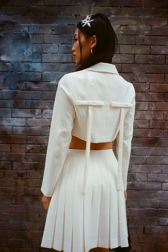 A model wears a bone colored skort and a Cropped tailored jacket in Bone mid weight suiting with long bows at back, chest pockets, and button closure against a brick wall