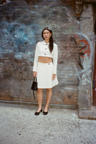 A model wears a bone colored skort and a Cropped tailored jacket in Bone mid weight suiting with long bows at back, chest pockets, and button closure against a brick wall