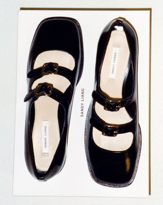 Black Mary Jane double strap shoes with a polished leather finish, featuring adjustable pin-buckle straps and a low heel against a white background