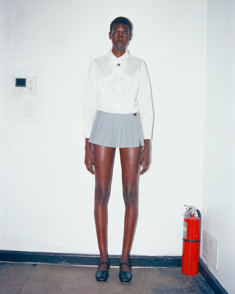 A woman wears a pleated micro skort in grey with built-in shorts and a button closure at the side with a white shirt tucked in against a white wall