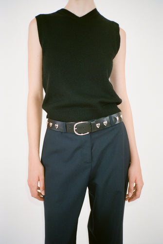 A person wearing a black leather belt with silver-toned heart studs and a silver buckle with black pants and a black sweater vest on a white background