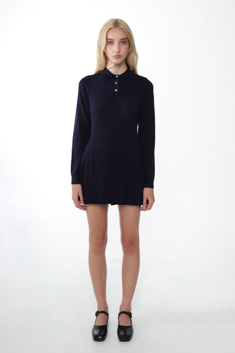 A woman wears a navy mini sweater dress with long sleeves and a polo neck. It features mother of pearl buttons in black shoes against a white background