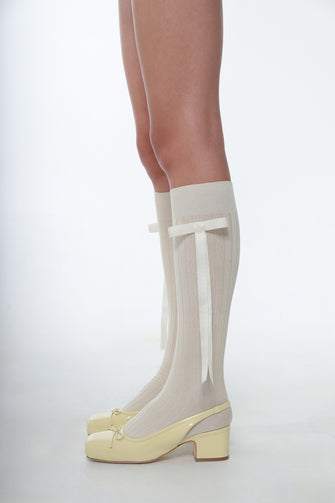 A person wearing knee high socks with long bows on them with a pair of yellow slingback heels in patent leather on a white background