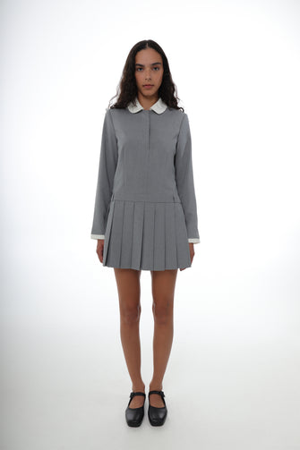 A woman wears a pleated mini dress in grey with a contrast white collar, long sleeves, and a side zipper with black shoes against a white background