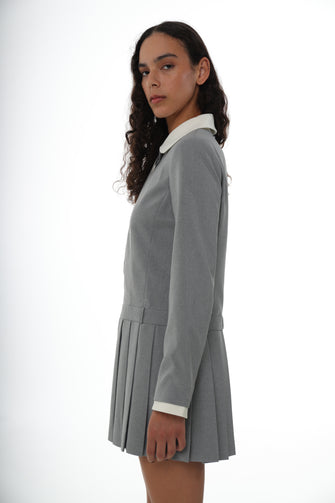 A woman wears a pleated mini dress in grey with a contrast white collar, long sleeves, and a side zipper against a white background