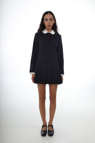 A woman wears a navy pleated mini dress with a contrast white collar and a slightly dropped waist, featuring a concealed zipper at the side and a boxy fit with black shoes against a white background