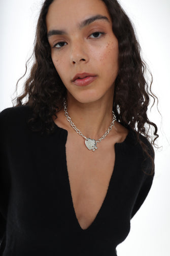 A girl wears a silver-colored oversized cable chain necklace with a heart tag pendant. The pendant is engraved with the text 'RETURN TO SANDY LIANG'. and a black v-neck sweater against a white background