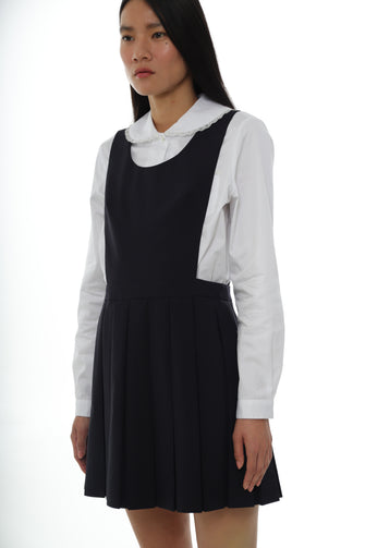 A woman wears a mini pleated navy pinafore dress with a scooped neckline over a white button down against a white background