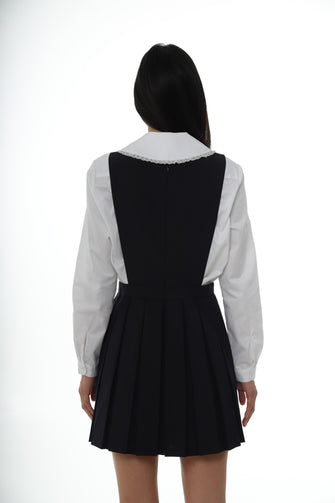 The back of a woman wearing a mini pleated navy pinafore dress with a scooped neckline over a white button down against a white background