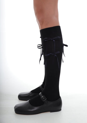 A pair of black knee-high socks with bows, worn with black shoes against a white background