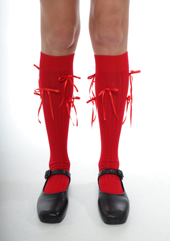 A pair of red knee-high socks with red bows, worn with black shoes against a white background