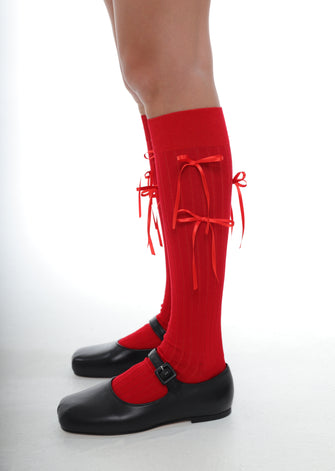 A pair of red knee-high socks with red bows, worn with black shoes against a white background