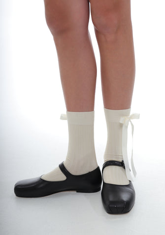 A pair of beige cotton blend crew socks with a ribbon bow on the side, worn with black ballet flats.