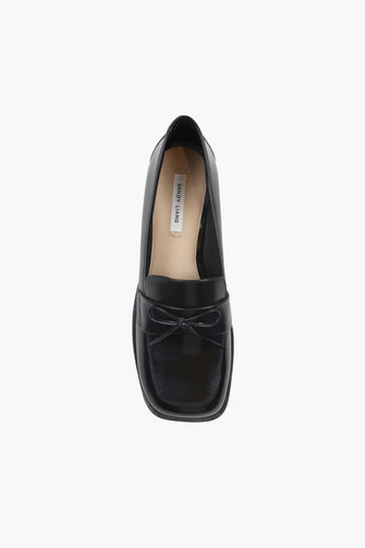 A top view of a black leather loafer with a bow detail on the vamp and a stacked leather heel on a white background