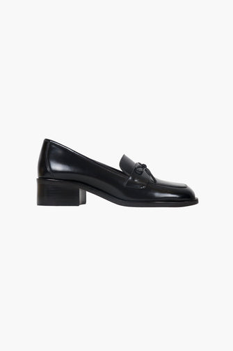 A side view of a black leather loafer with a bow detail on the vamp and a stacked leather heel on a white background