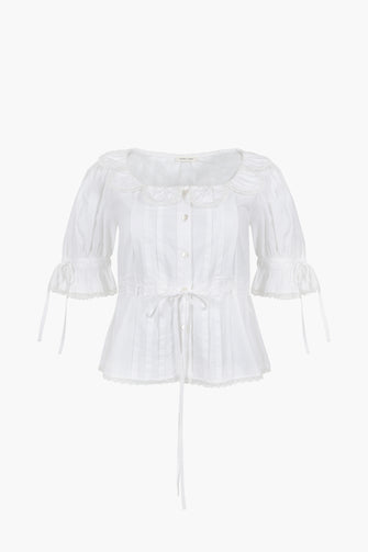 A white button down shirt with a ruffle collar and bows on the sleeves and waist against a white background