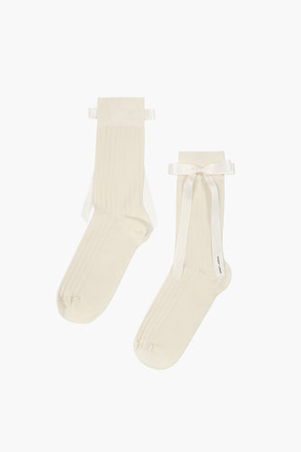 A pair of cream-colored socks with a ribbon bow on the side, made from cotton blend fabric against a white background