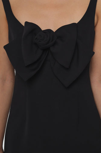 A woman wears a tailored mini dress in black with oversized bow at front against a white wall