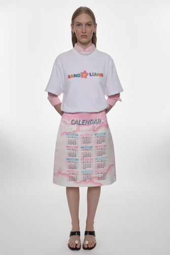 Person wearing a white oversized t-shirt with colorful text and a pink skirt with calendar print on a white background
