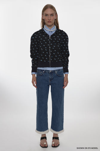 Person wearing a black patterned jacket, light blue shirt, and blue jeans on a white background
