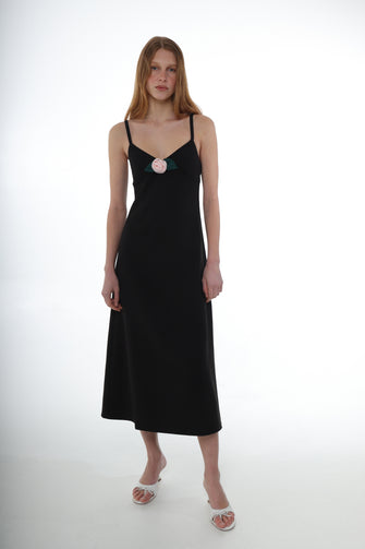 A woman wears a A midi length black dress with thin straps, a flower detail at the chest, and an invisible side zip. The dress has a tailored silhouette with a slight flare at the hem with white heels against a white background