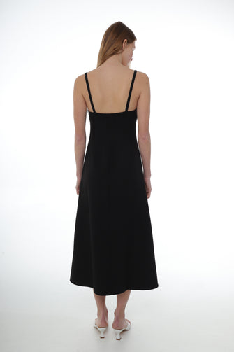 A woman wears a A midi length black dress with thin straps, a flower detail at the chest, and an invisible side zip. The dress has a tailored silhouette with a slight flare at the hem with white heels against a white background