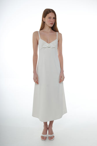 A woman wears a midi-length ivory dress with thin straps, a flower detail at the chest, and a side zipper with white heels against a white background