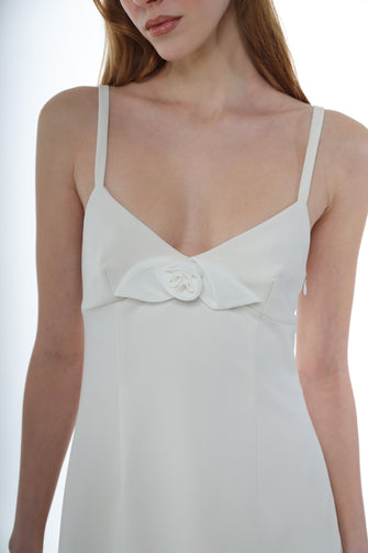 A woman wears a midi-length ivory dress with thin straps, a flower detail at the chest, and a side zipper against a white background