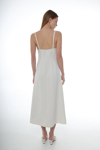 A woman wears a midi-length ivory dress with thin straps, a flower detail at the chest, and a side zipper with white heels against a white background
