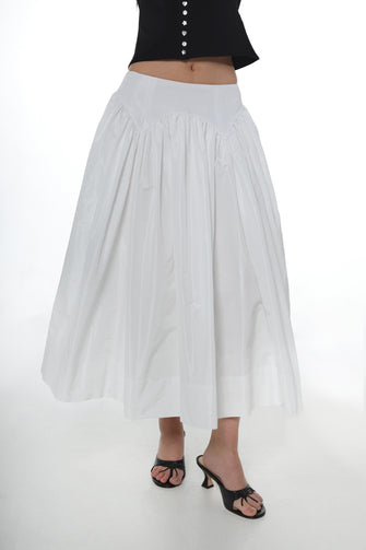 A woman wears a full length white skirt with a pleated design with a black cardigan and black heels against a white background