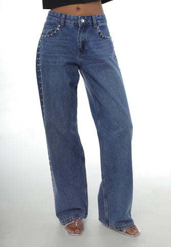 A woman in pair of blue denim jeans with rhinestone rivets at the sides against a white background