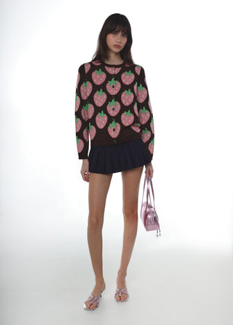 Woman wearing a brown sweater with pink strawberry pattern and brown skirt holding a pink bag on a white background