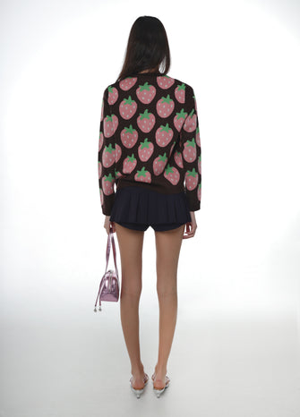 Woman wearing a brown sweater with pink strawberry pattern and brown skirt holding a pink bag on a white background
