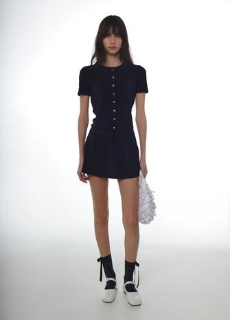 Woman wearing a navy button down jersey shirt with a navy jersey skort with navy socks, white mary jane shoes and a white ruffled bag on a white background