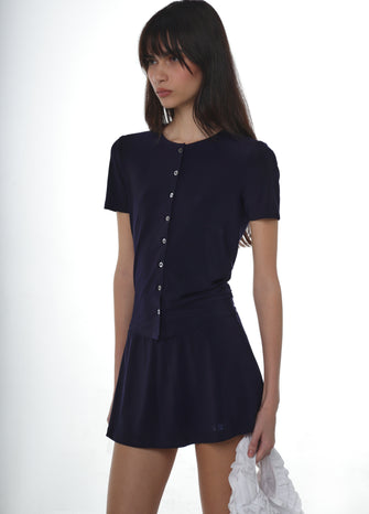 Woman wearing a navy button down jersey shirt with a navy jersey skort with a white ruffled bag on a white background