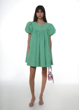 Woman wearing a green checkered babydoll dress ,silver shoes, and a pink handbag on a white background