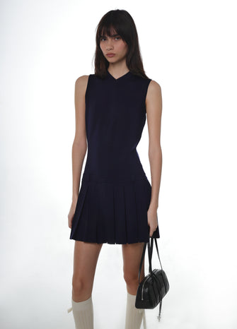 Woman wearing a navy sleeveless knit dress holding a black handbag with white knee socks against a white background