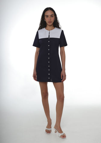 Woman wearing a navy cotton dress with button details and an oversized white sailor collar and white heels on a white background