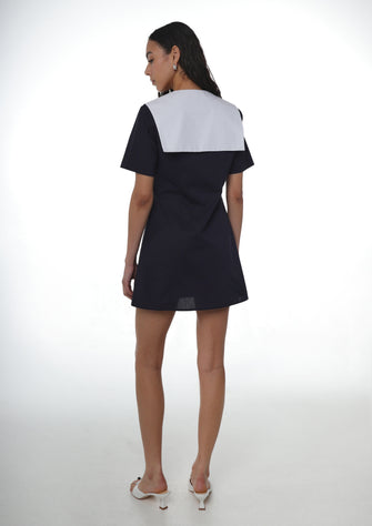 Woman wearing navy cotton dress with button details and an oversized white sailor collar and white heels on a white background