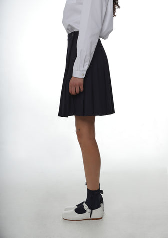 A close up of someone wearing a white peter pan collared shirt tucked into a navy pleated skirt with white shoes and navy socks against a white background