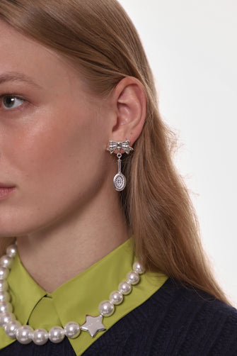 Close-up of a person wearing a pearl necklace and silver bow and hairbrush earrings with a plain background