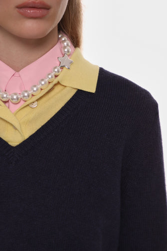 Close-up of a person wearing a navy sweater with a yellow collar, pink shirt, and pearl necklace.
