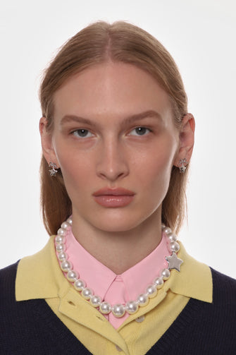 Woman wearing a pearl necklace, pink shirt, and yellow cardigan with silver earrings on a white background