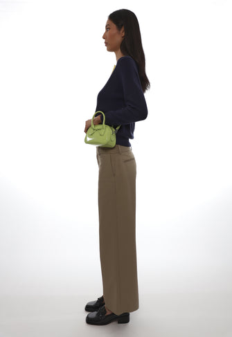 Woman holding a green handbag wearing tan pants and a navy sweater against a white background