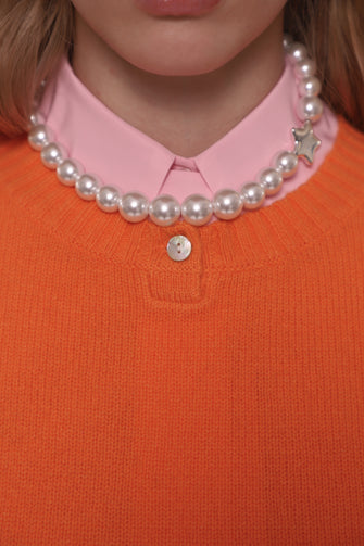 Person wearing an orange sweater and pink shirt with a pearl necklace featuring a star pendant.