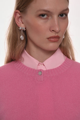 Woman wearing a pink sweater and shirt with earrings against a white background