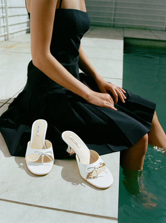 A woman sits on the edge of a pool in a black dress with white shoes next to her