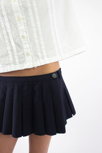 A closeup of a person wearing pleated dark navy micro skort with built-in shorts and a button closure at the side with a white button down shirt against a white background.